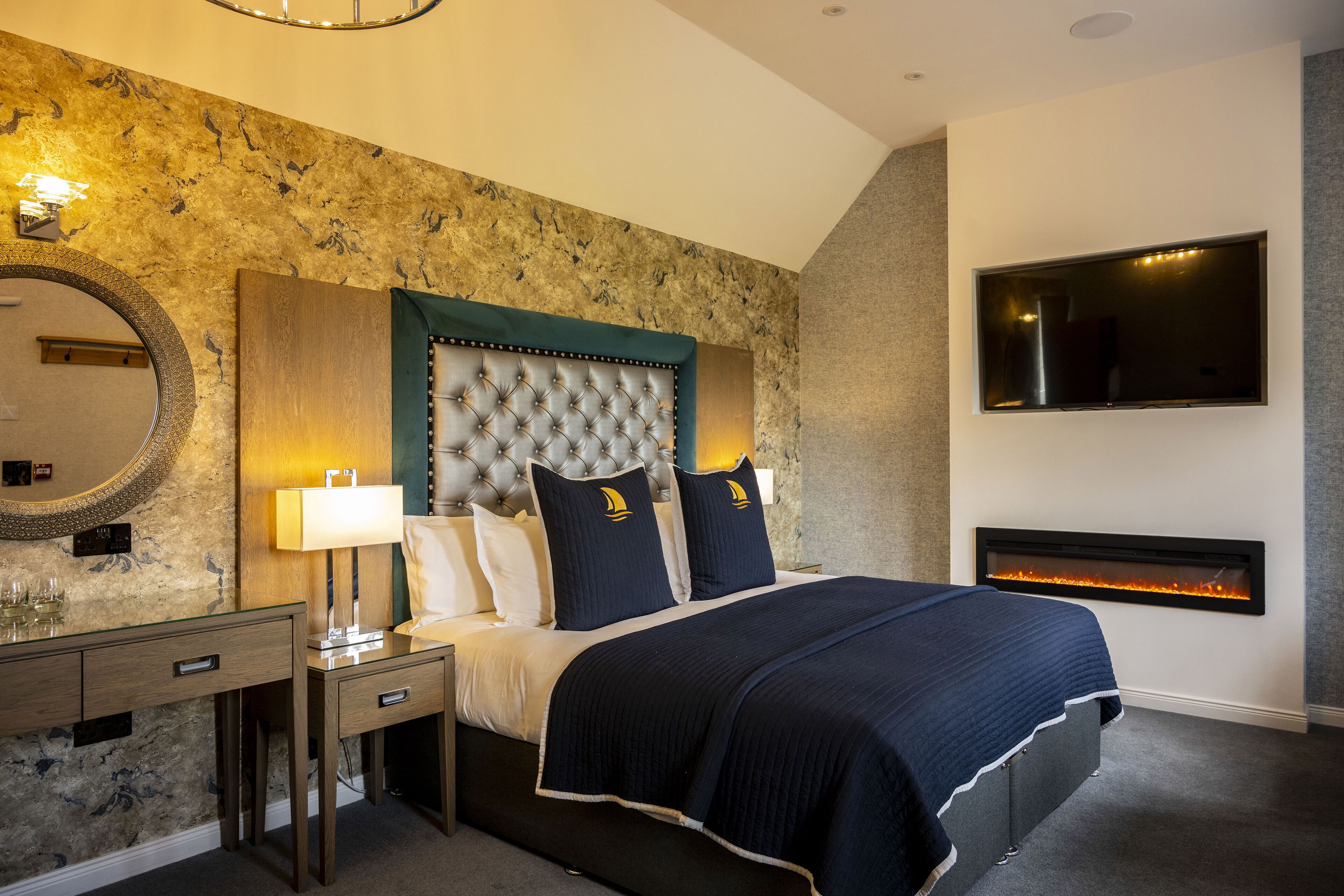 A modern bedroom with a tufted headboard, a fireplace, a large mirror, and a flat-screen TV.