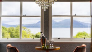 Una habitación con vista a un lago y montañas, una mesa con una botella y copas, y dos sillones a cuadros.