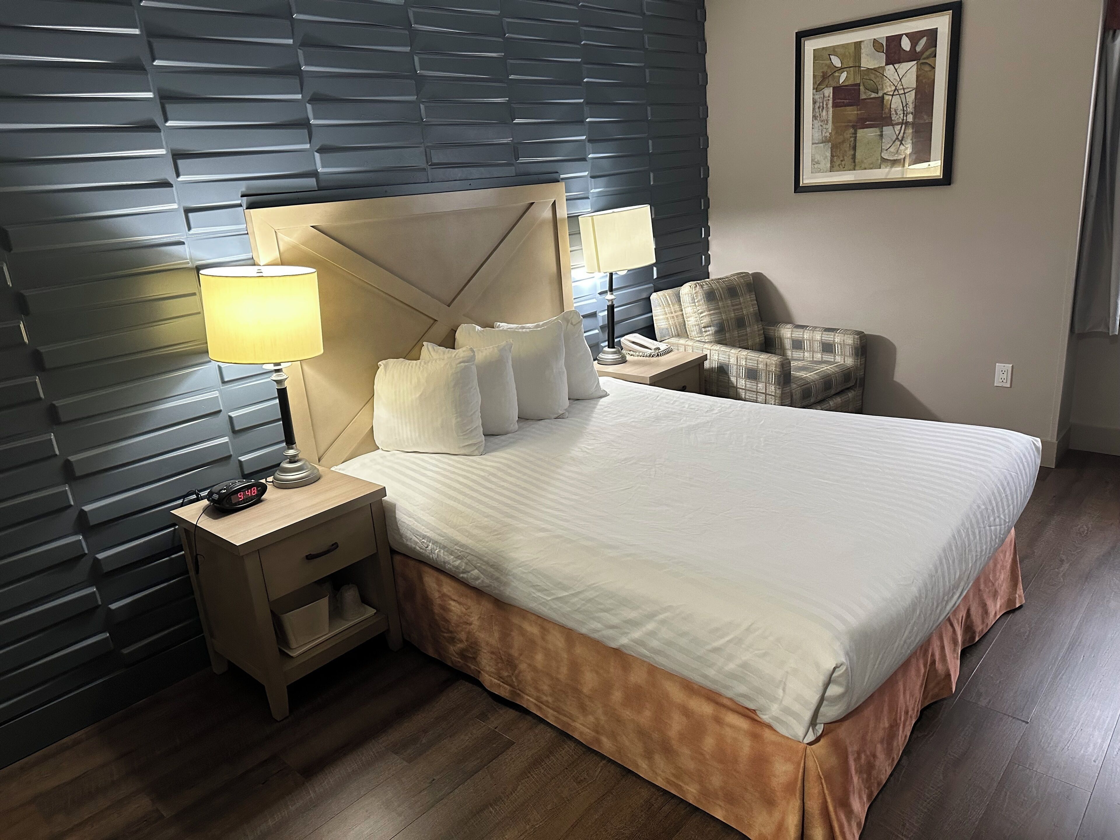 A hotel room with a large bed, a nightstand with a lamp, a chair, and a wall-mounted artwork.