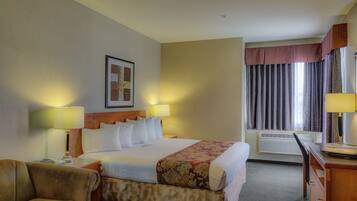Quarto de hotel com uma cama grande, duas luminárias de cabeceira, uma escrivaninha com cadeira, um sofá e uma janela com cortinas.