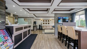 A hotel lobby with a reception desk, a seating area with a sofa and chairs, a television mounted on the wall, and a window with a view of greenery.