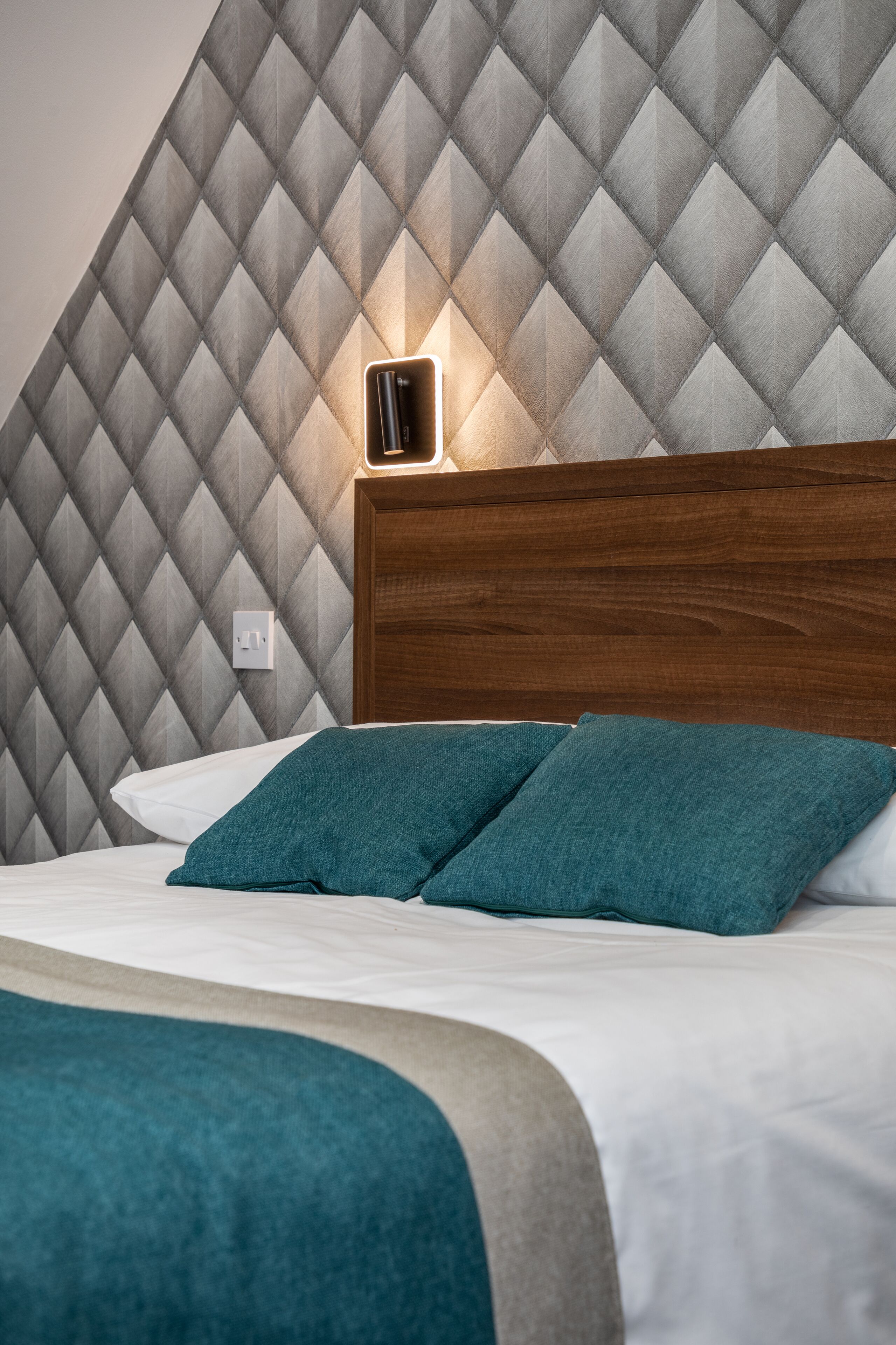 A neatly made bed with teal pillows and a wooden headboard against a diamond-patterned wall.