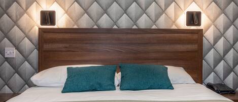 Una cama bien hecha, con un cabecero de madera, dos almohadas color turquesa y una blanca. Hay dos lámparas de noche y un teléfono negro en la mesita.