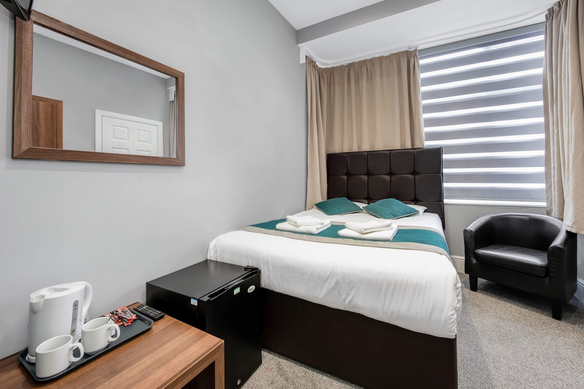 A neatly arranged hotel room with a bed, a chair, a desk with a coffee maker, and a mirror.