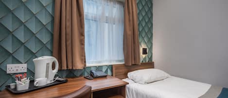 A hotel room with a wooden desk, a bed, a window with curtains, a wall with a geometric pattern, and a nightstand with a lamp.