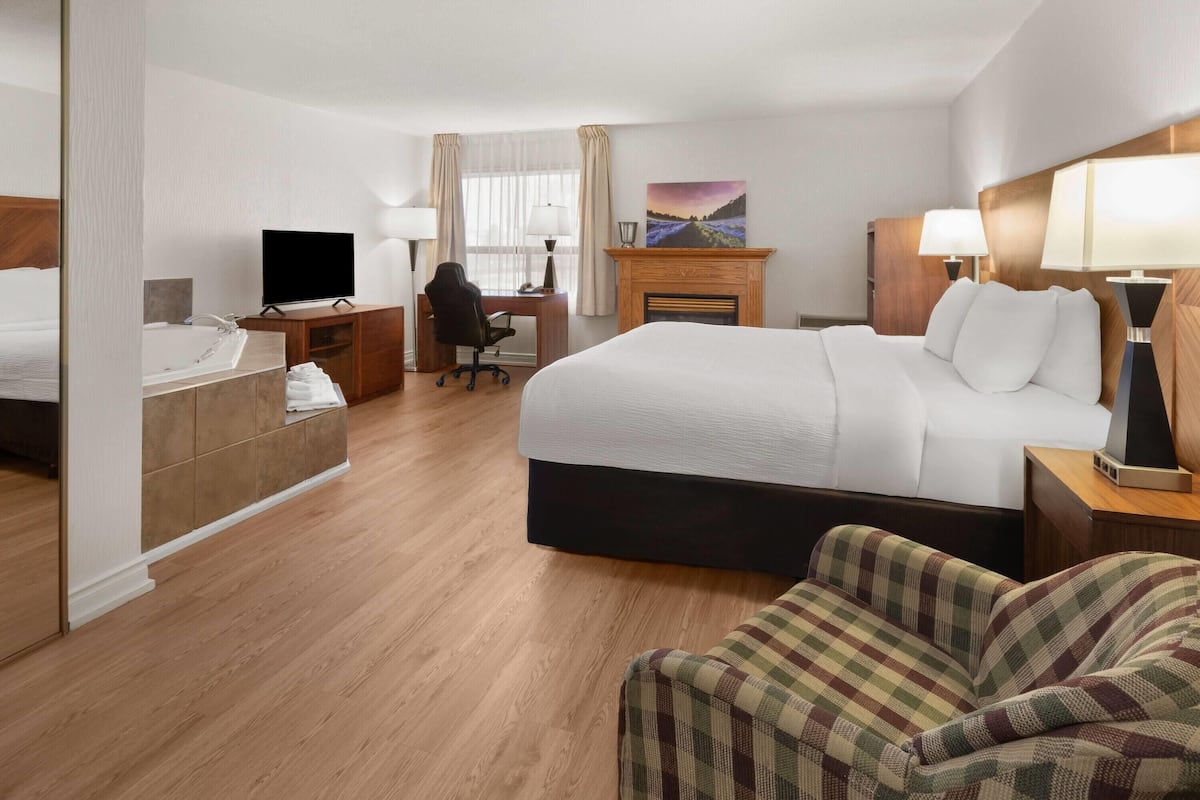 A hotel room with a large bed, a desk with a chair, a television, a fireplace, and a view of a landscape painting.
