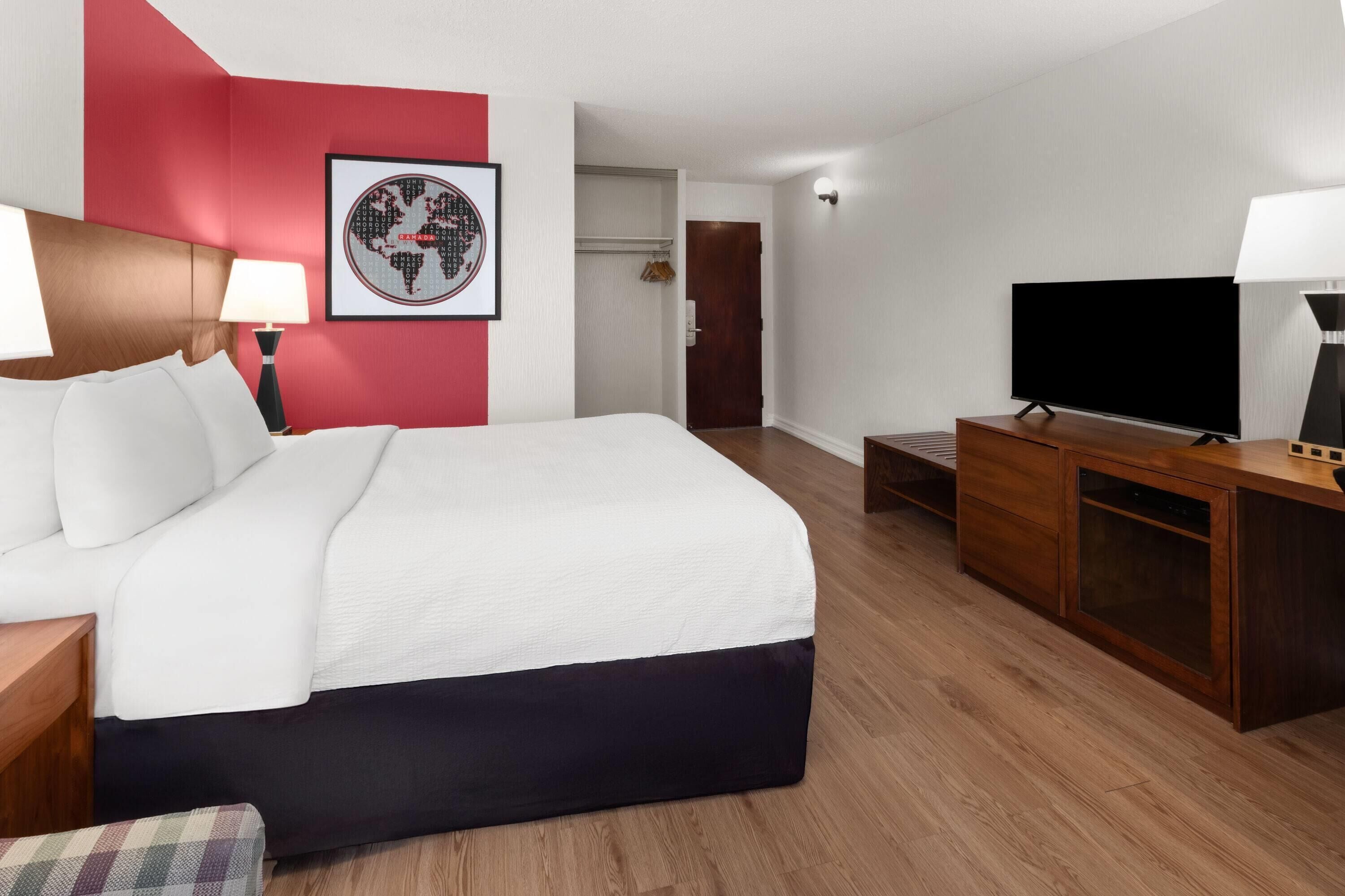 A hotel room with a bed, a TV, a desk, a lamp, and a framed picture on the wall.
