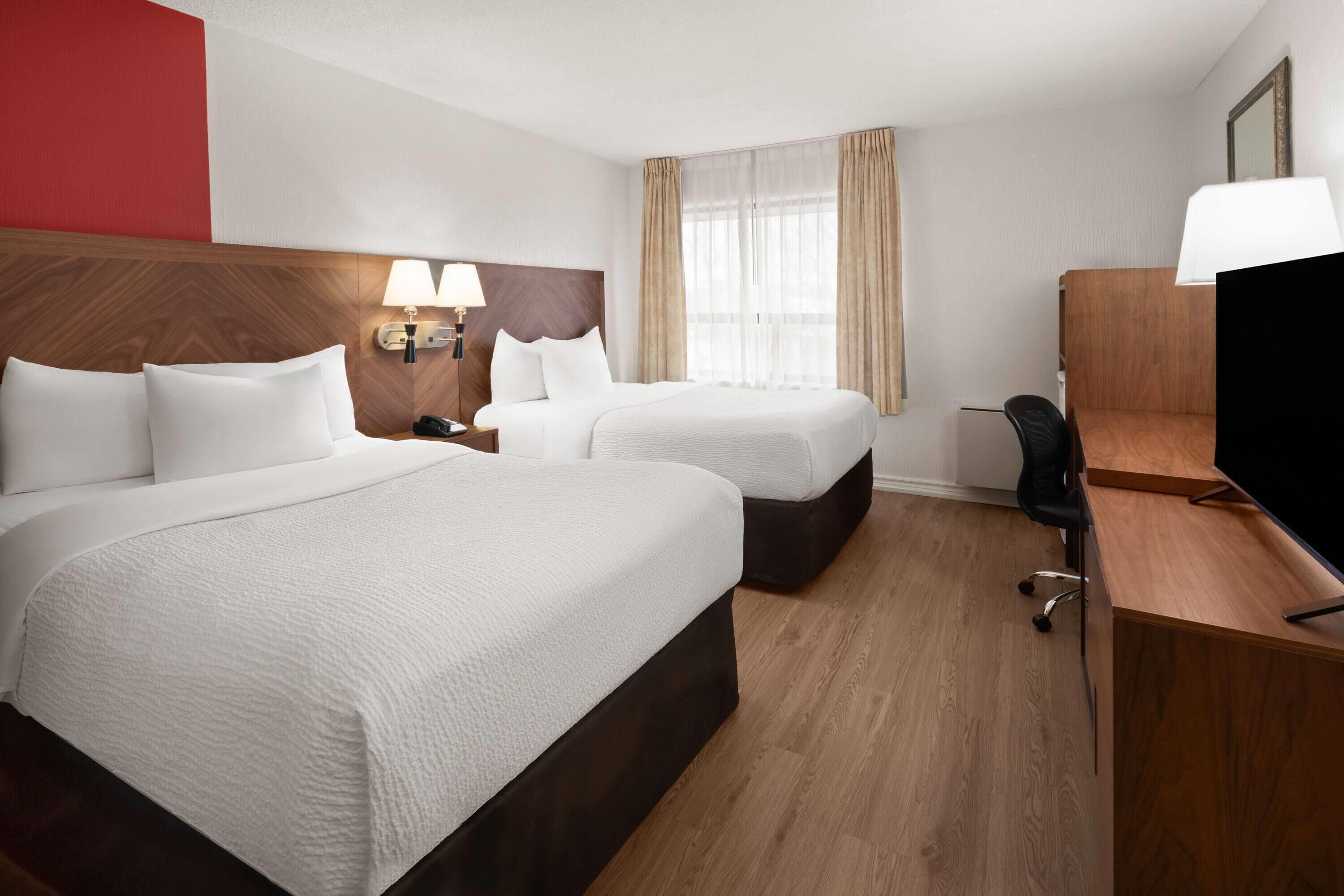 A hotel room with two beds, a desk, a chair, a TV, and a lamp.