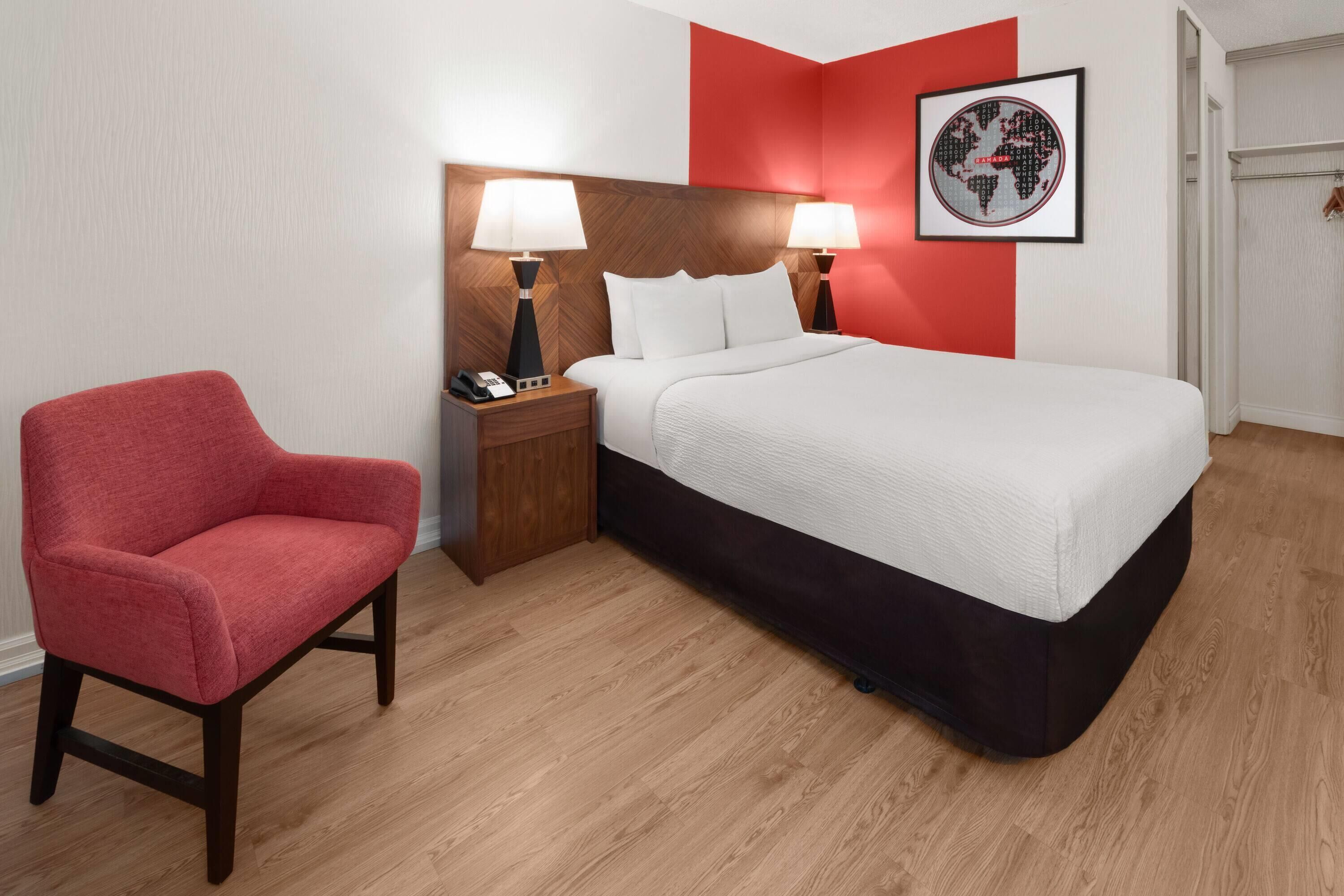 Una habitación de hotel con una cama, dos lámparas de noche, una silla roja, una mesita de noche de madera y un cuadro en la pared.