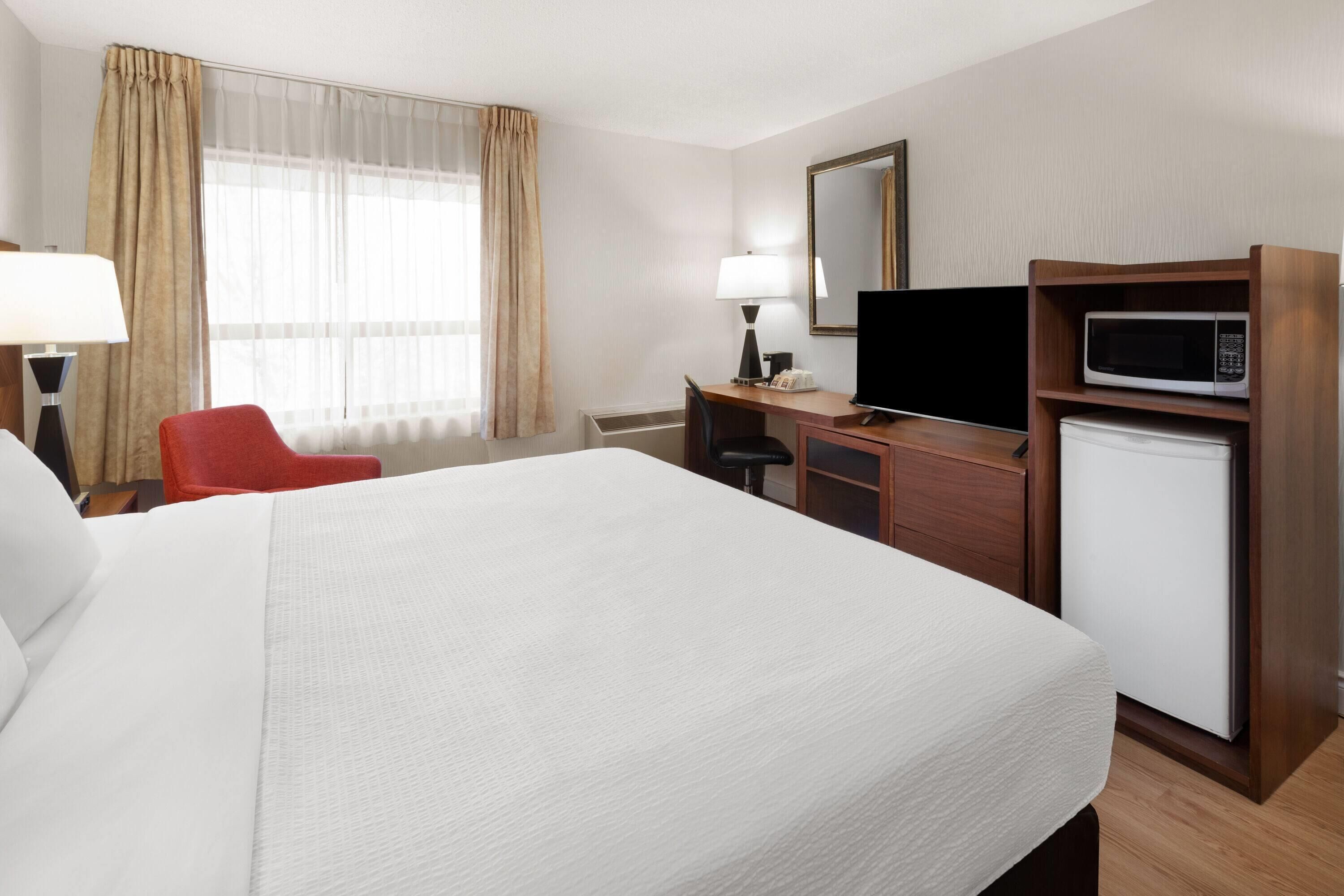 Quarto de hotel com uma cama grande, uma mesa com cadeira, televisão, micro-ondas e janela com cortinas.