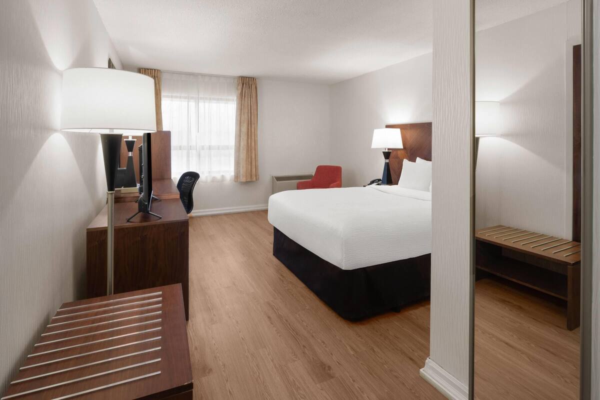 A hotel room with a bed, desk, chair, bedside table, lamp, and a small seating area.