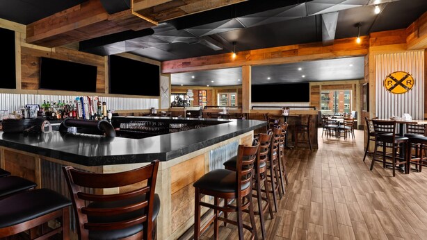 Una zona de bar con una isla central, taburetes de madera y un área de asientos con mesas y sillas.