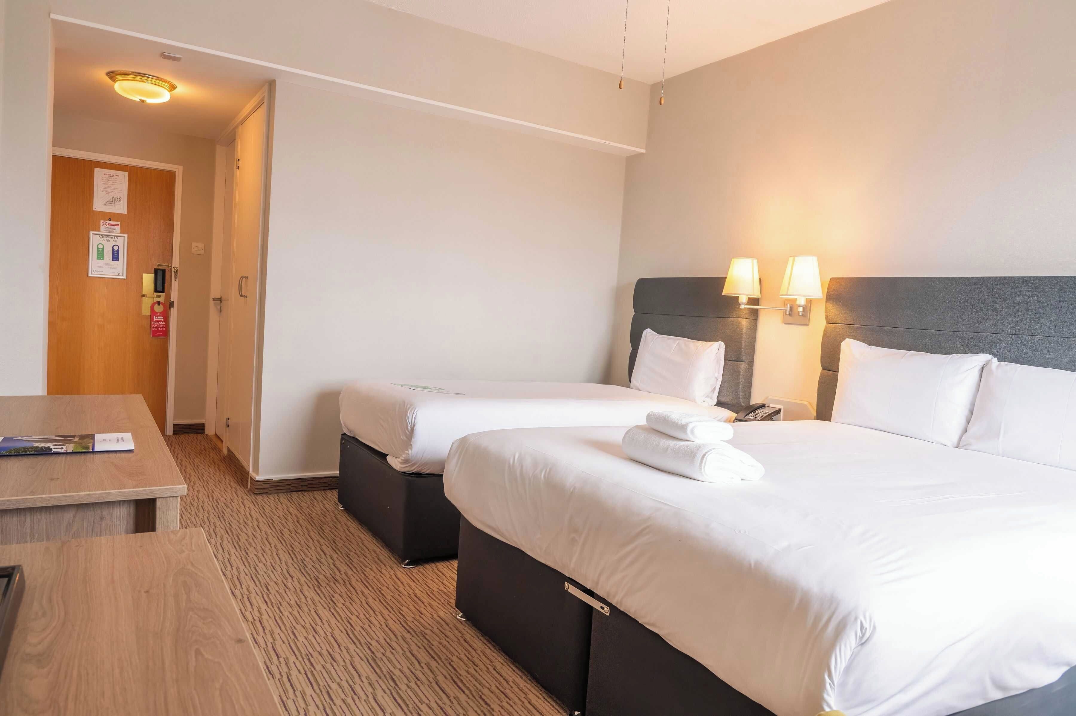A hotel room with two beds, a desk, and a chair. There are two wall-mounted lamps and a ceiling light.