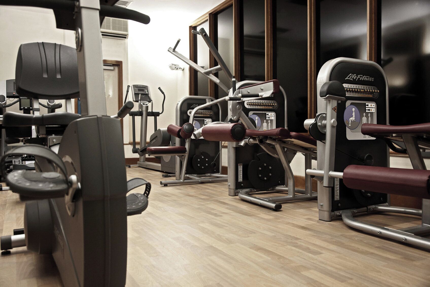 A gym with various exercise machines, including treadmills and weight machines, arranged in a spacious room with wooden flooring.