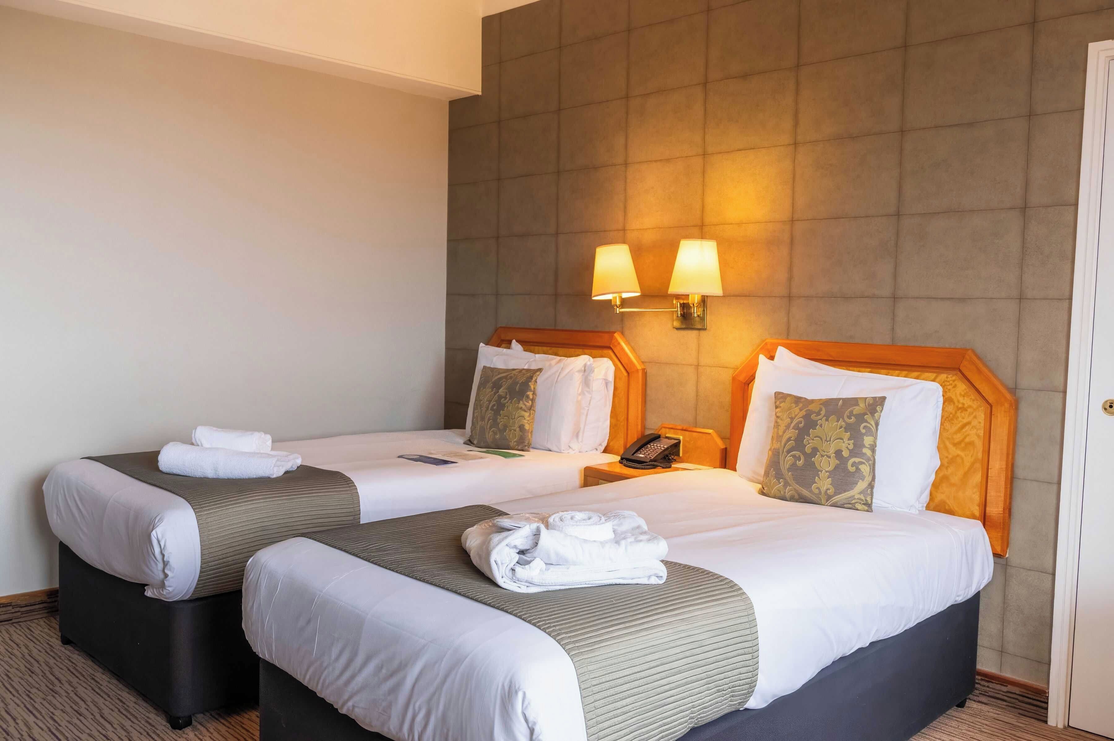 Een hotelkamer met twee bedden, elk voorzien van een kussen, een nachtkastje met een telefoon en een wandlamp.