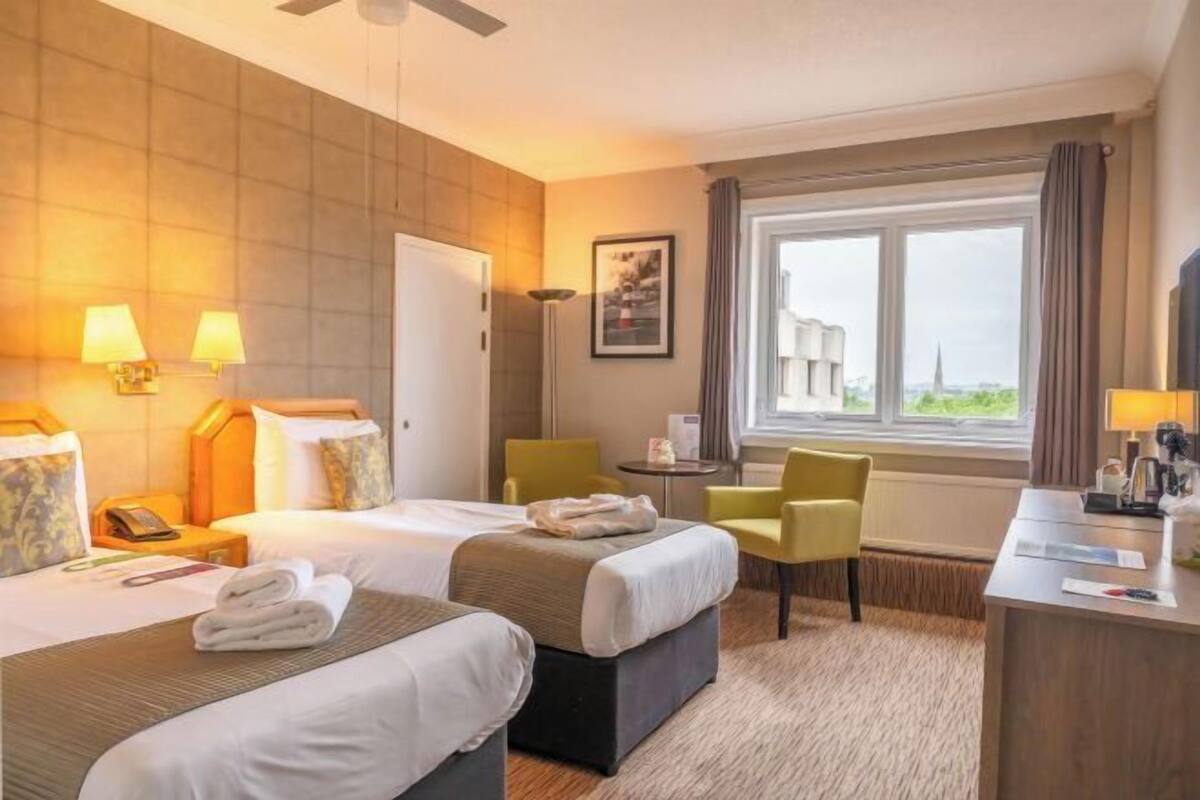 Ein Hotelzimmer mit zwei Betten, einem Schreibtisch, einem Stuhl, einem Fenster mit Blick und einem Deckenventilator.