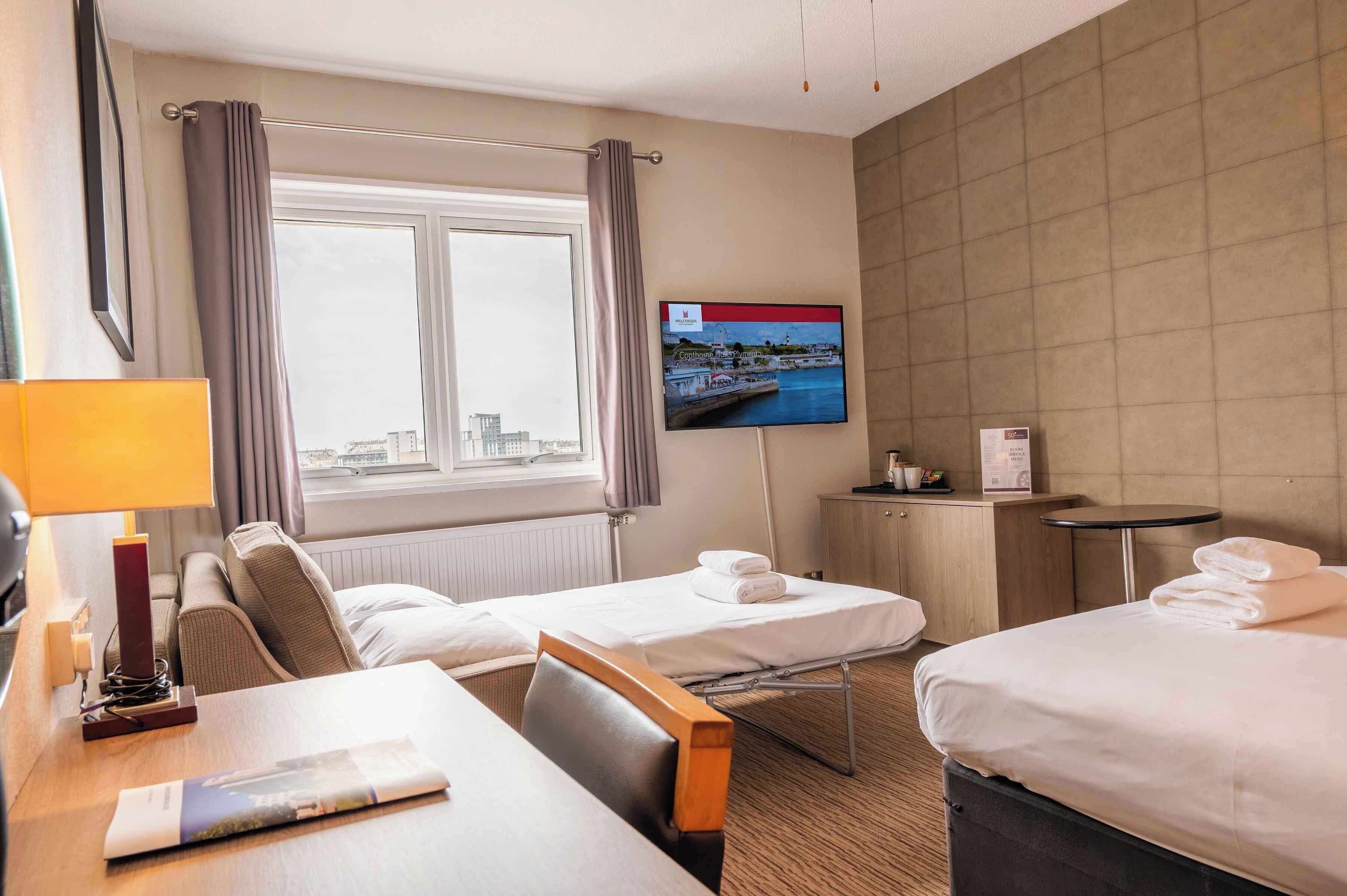 A hotel room with two beds, a TV on the wall, a desk with a lamp, and a view of the city through the window.