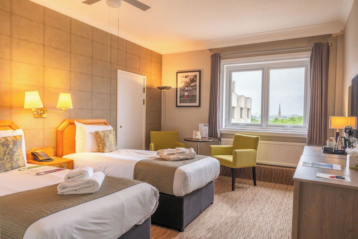 Ein Hotelzimmer mit zwei Betten, einem Schreibtisch, einem Stuhl, einem Fenster mit Blick und einem Deckenventilator.