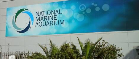The exterior of the National Marine Aquarium with a large blue and white sign, a gated entrance, and some greenery in front.