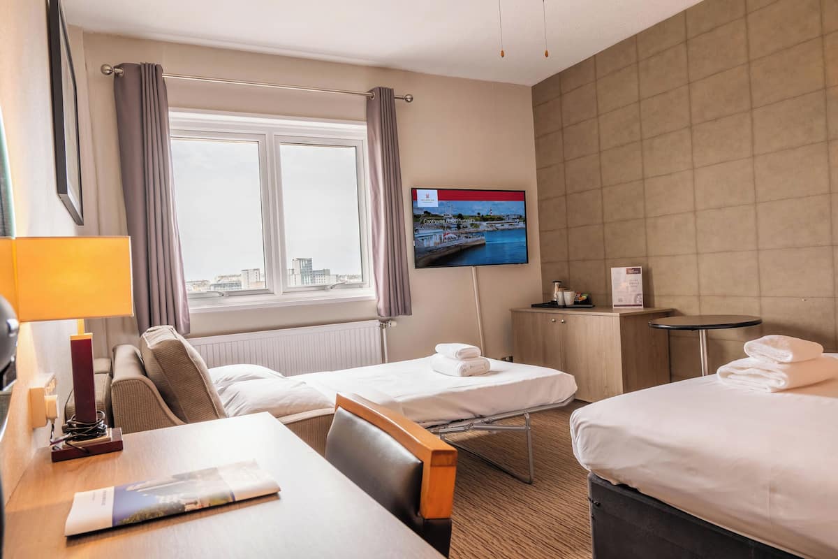 Ein Hotelzimmer mit zwei Betten, einem an der Wand montierten Fernseher, einem Schreibtisch mit Lampe und Blick auf die Stadt durch das Fenster.