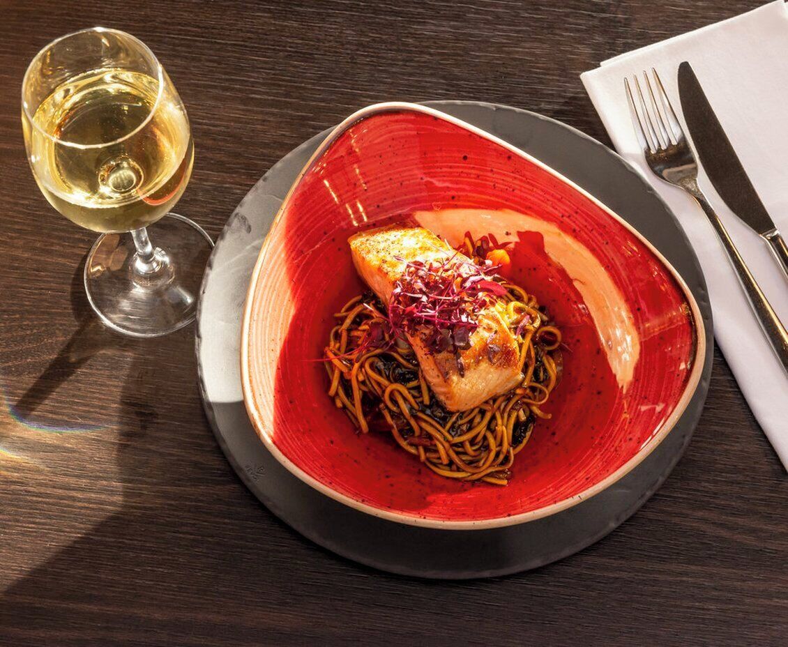 A plate of pasta with a piece of salmon, a glass of white wine, and cutlery on a dark wooden table.