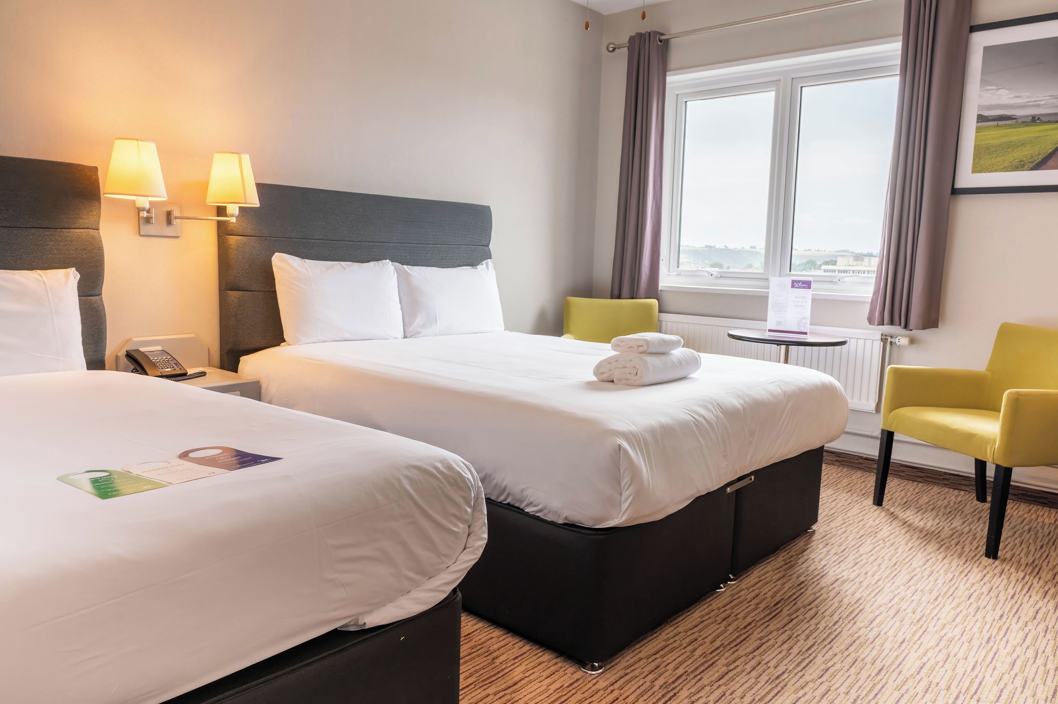 A hotel room with two beds, a small table, a chair, and a window with a view.