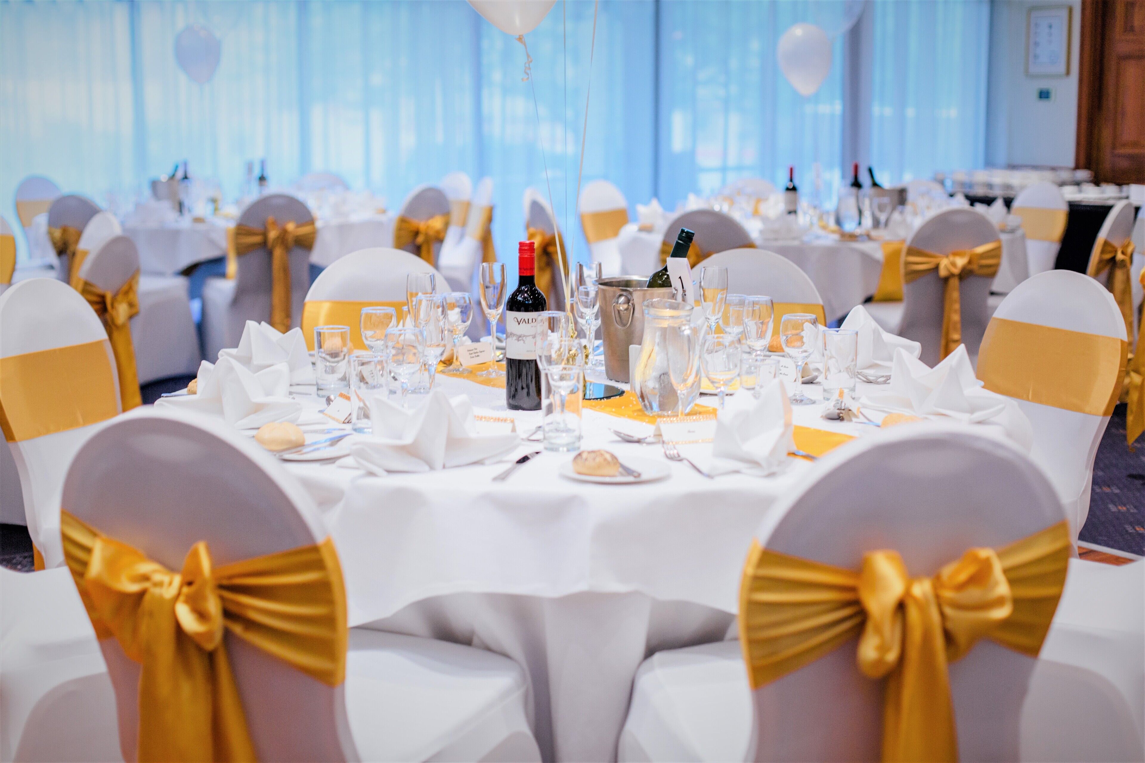 A banquet hall with round tables set for a formal event, featuring white tablecloths and gold sashes on chairs.