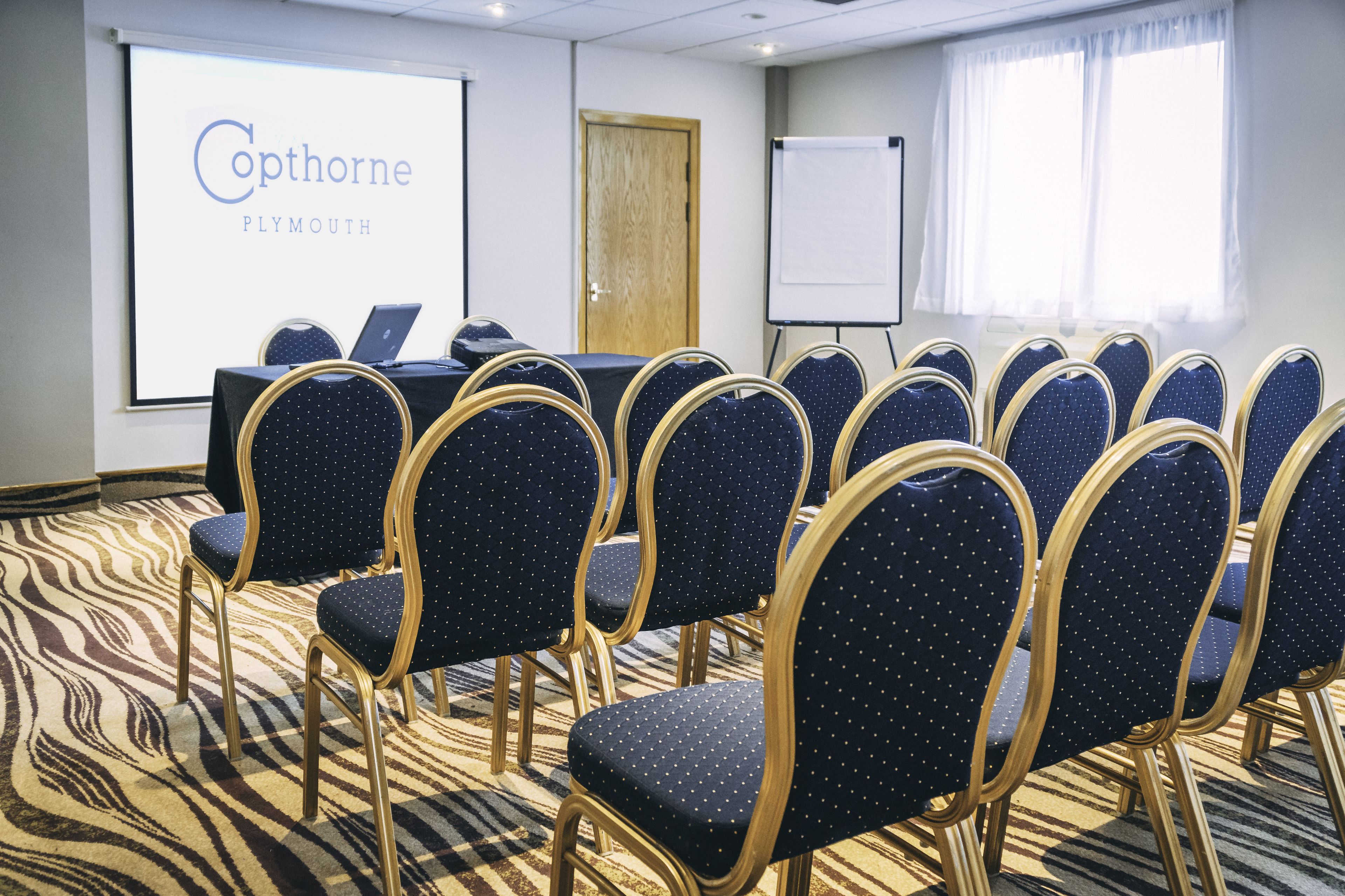 A conference room with a presentation screen displaying "Coptorne Plymouth," a table with laptops, and rows of chairs with a zebra-patterned carpet.