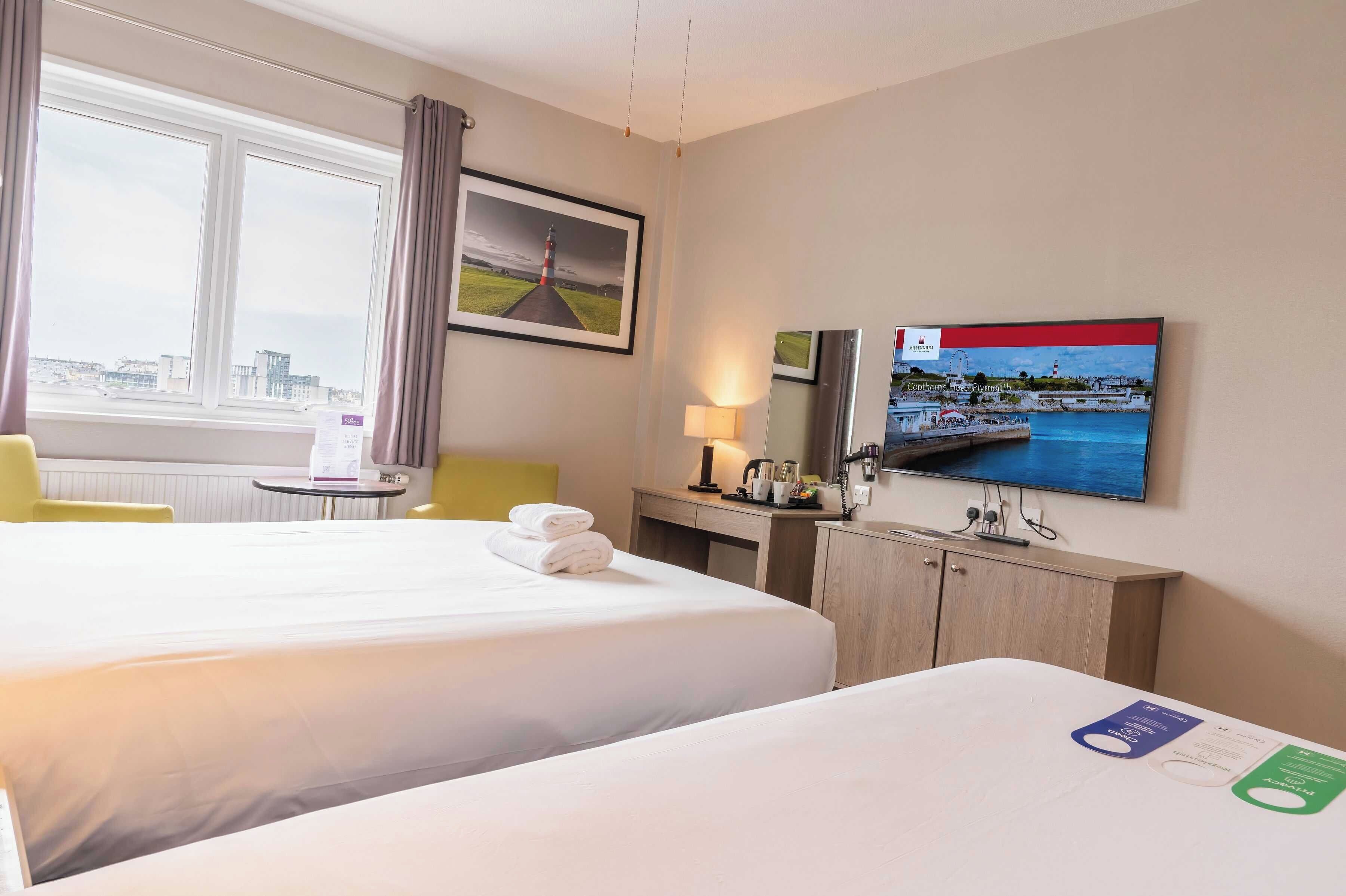 A hotel room with two beds, a desk, a chair, a TV, and a window with a view of the city.