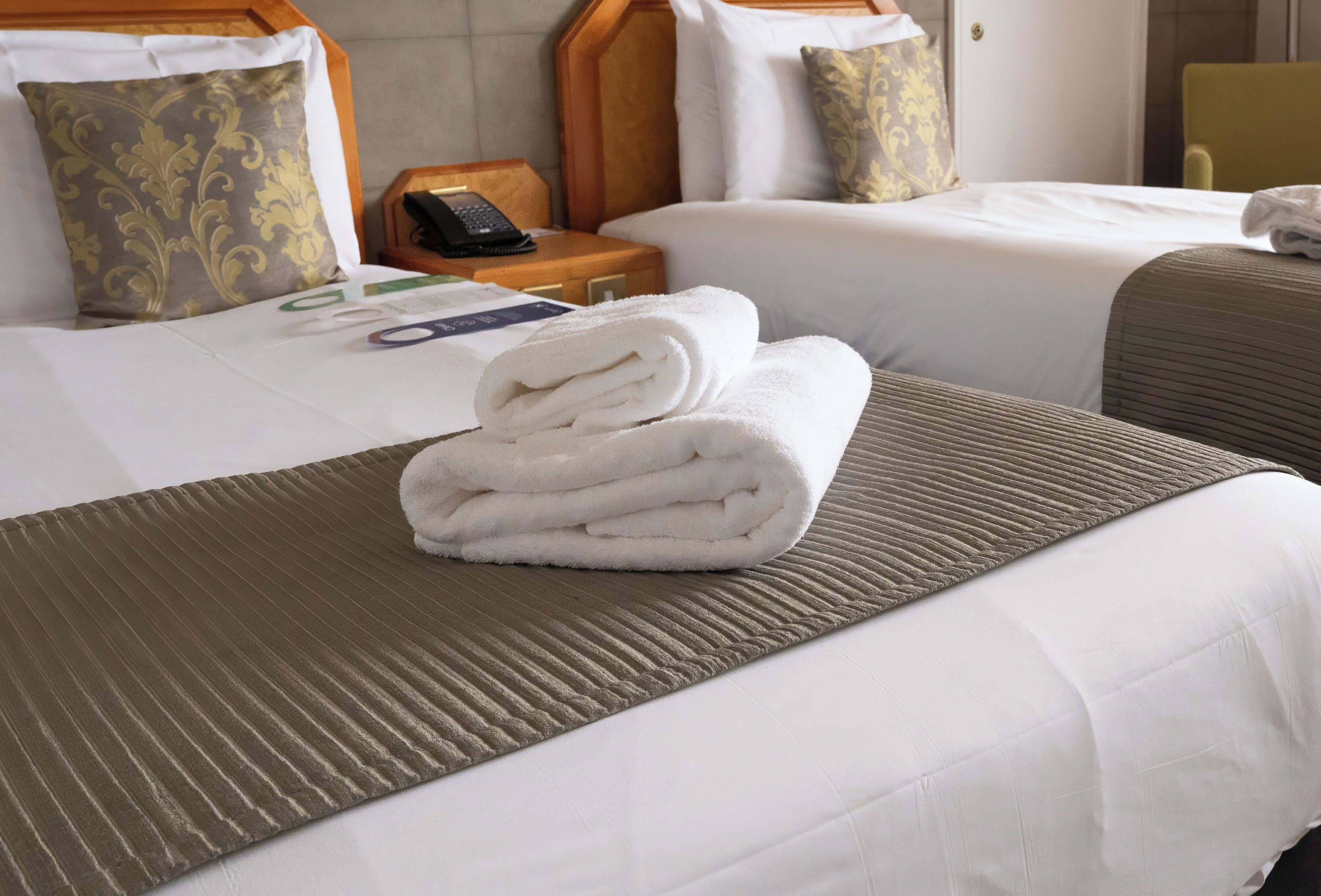 A hotel room with two beds, a wooden nightstand with a phone, and a folded towel on the bed.