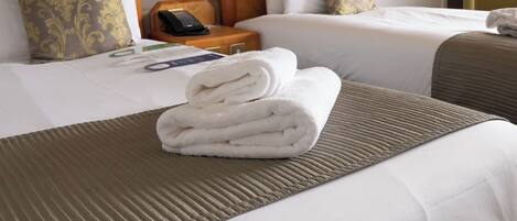 A hotel room with two beds, a wooden nightstand with a phone, and a folded towel on the bed.