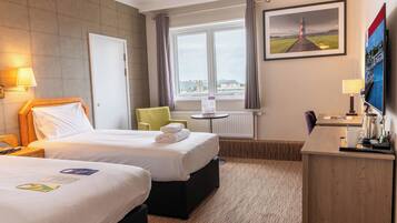 A hotel room with two beds, a desk, a chair, a window with a view, and a painting on the wall.