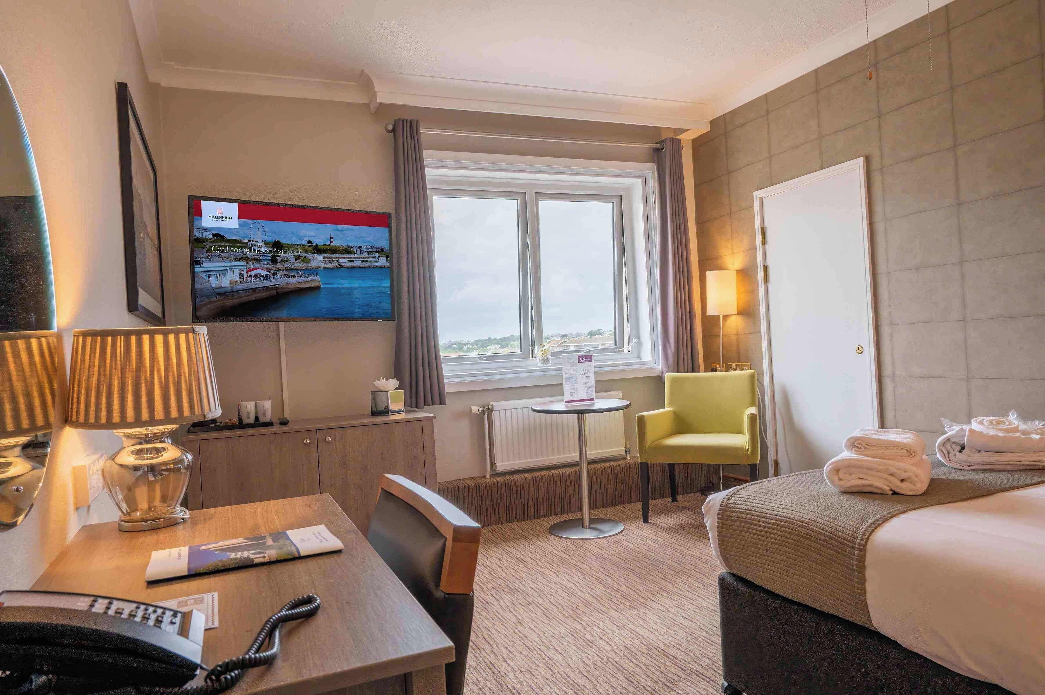 A hotel room with a bed, a desk with a lamp, a chair, a TV on the wall, and a window with a view.