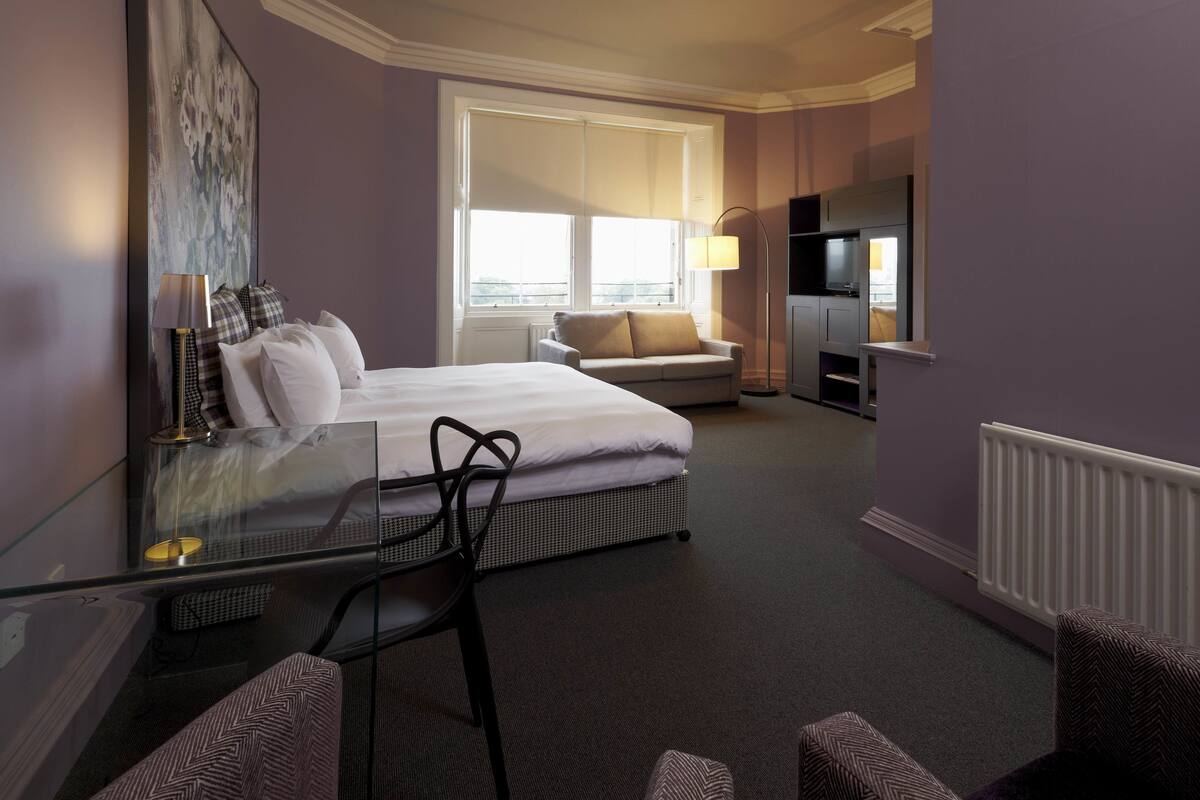 Habitación de hotel con cama, escritorio de cristal, silla, sofá, televisor y ventana con vistas.