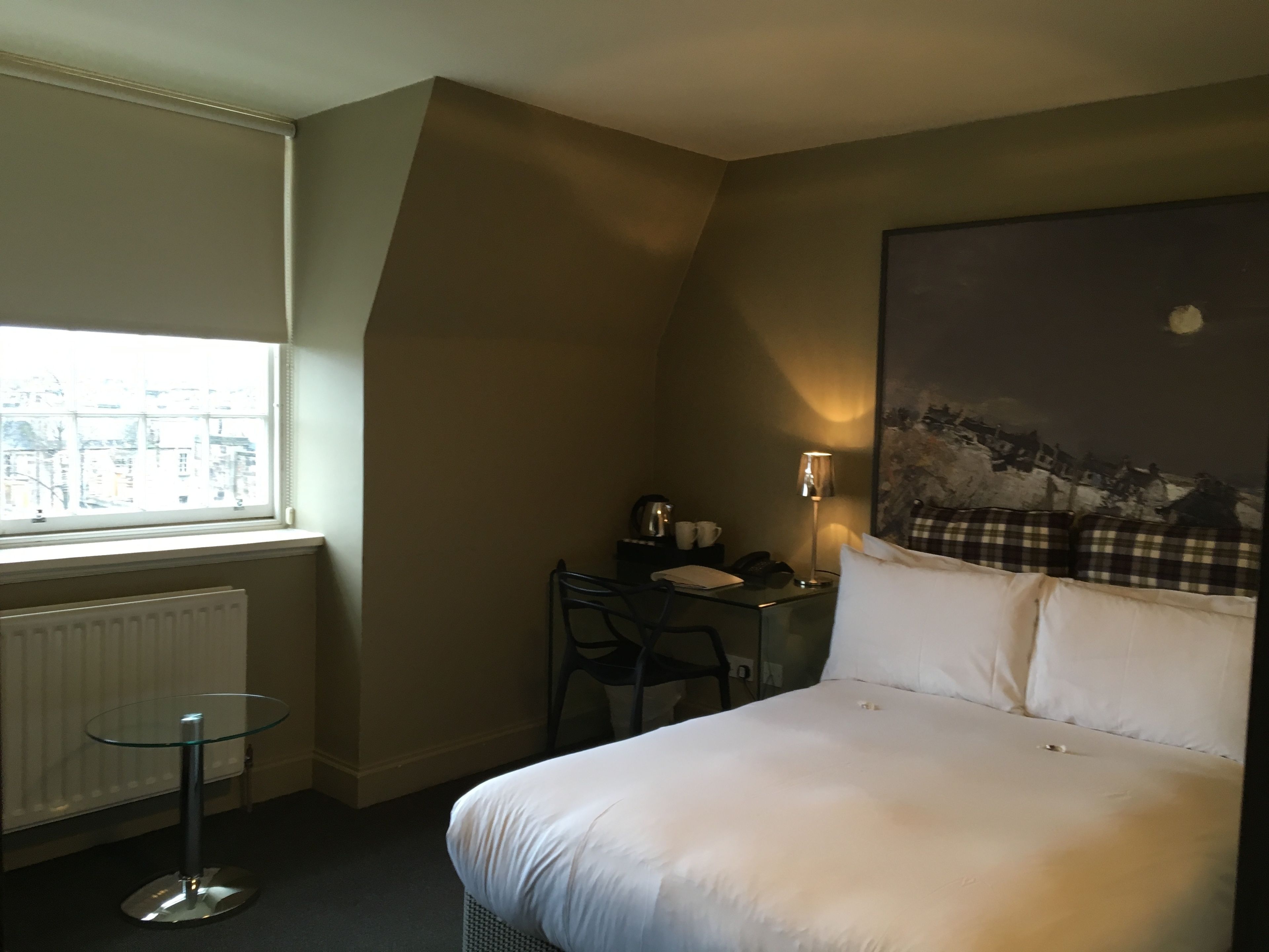 Habitación de hotel con cama, escritorio, silla, lámpara y ventana con vista a edificios.