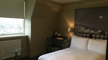 Une chambre d’hôtel avec un lit, un bureau et une chaise, une lampe et une fenêtre donnant sur des immeubles.