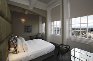 A bedroom with a large bed, a chair, a table, and a view of the city through large windows.