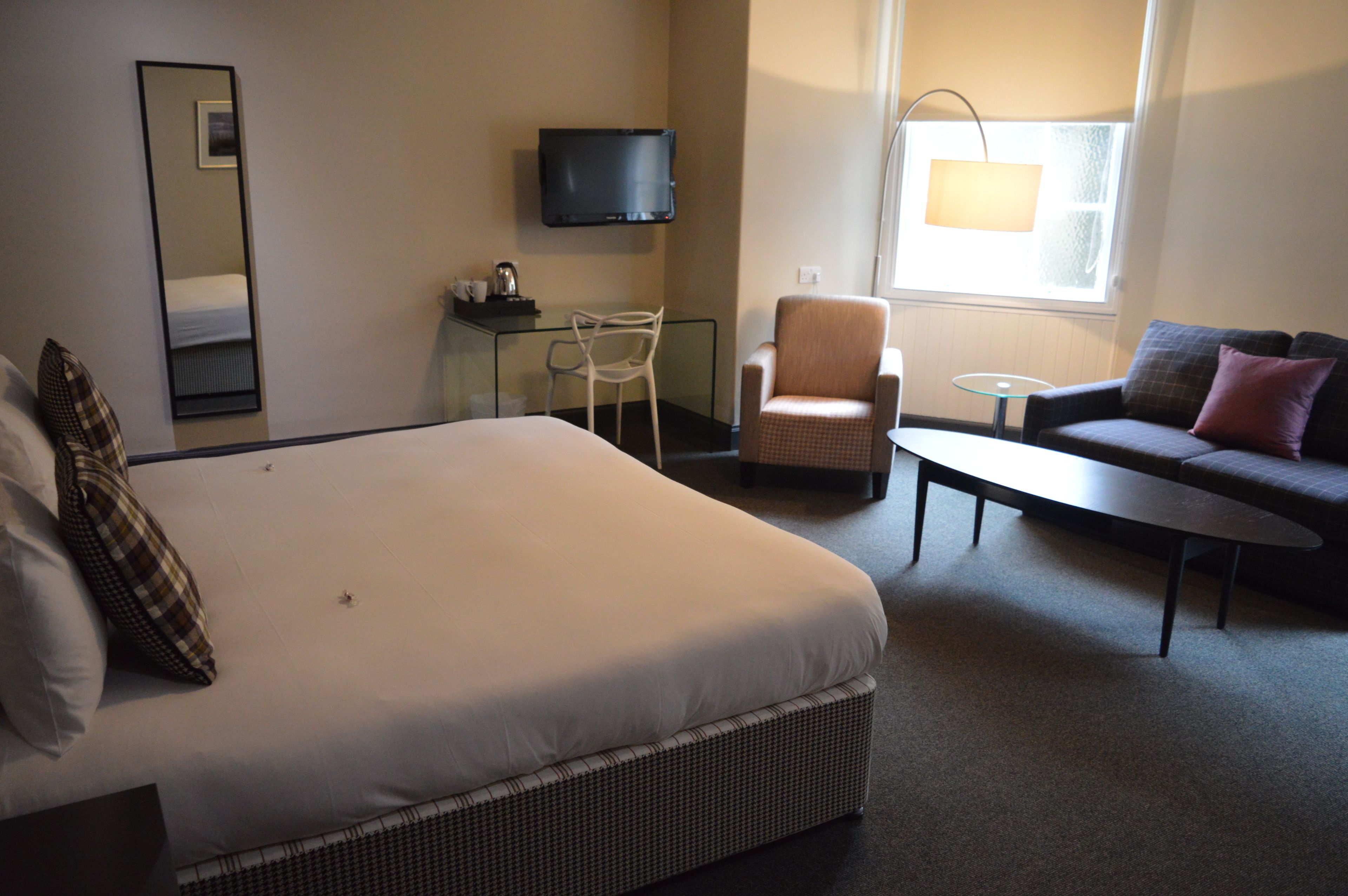 Une chambre d’hôtel avec un grand lit, un canapé, un fauteuil, une petite table, une télévision et une fenêtre avec une lampe.
