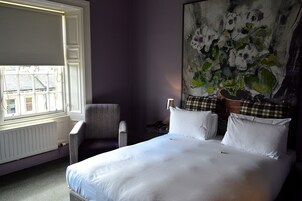 A hotel room with a bed, a chair, a radiator, and a window with blinds.