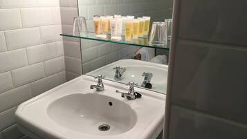 A bathroom with a white sink, two faucets, and a glass shelf above the sink holding toiletries.