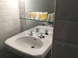 A bathroom with a white sink, two faucets, and a glass shelf above the sink holding toiletries.