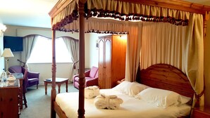A four-poster bed with white linens and towels, a wooden nightstand, a desk with a lamp, a purple armchair, and a wardrobe. - Skylark Hotel (Southend-on-Sea)