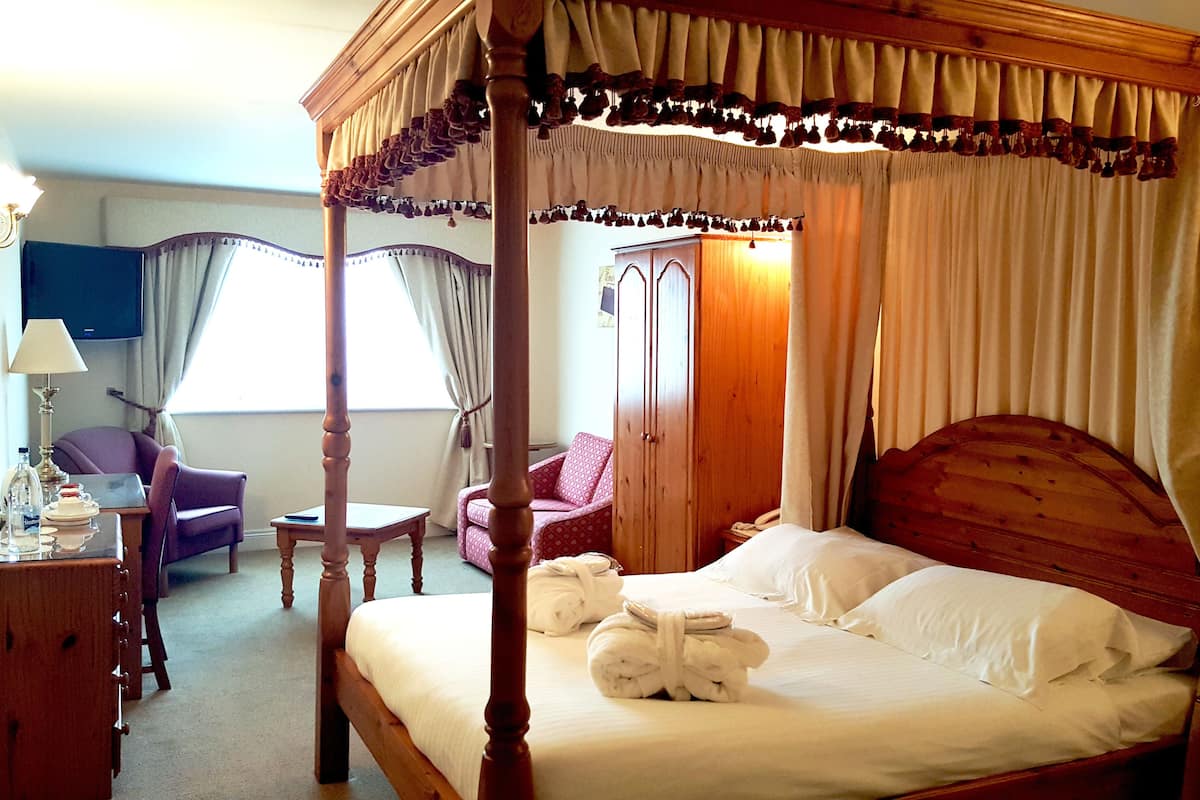 A four-poster bed with white linens and towels, a wooden nightstand, a desk with a lamp, a purple armchair, and a wardrobe.