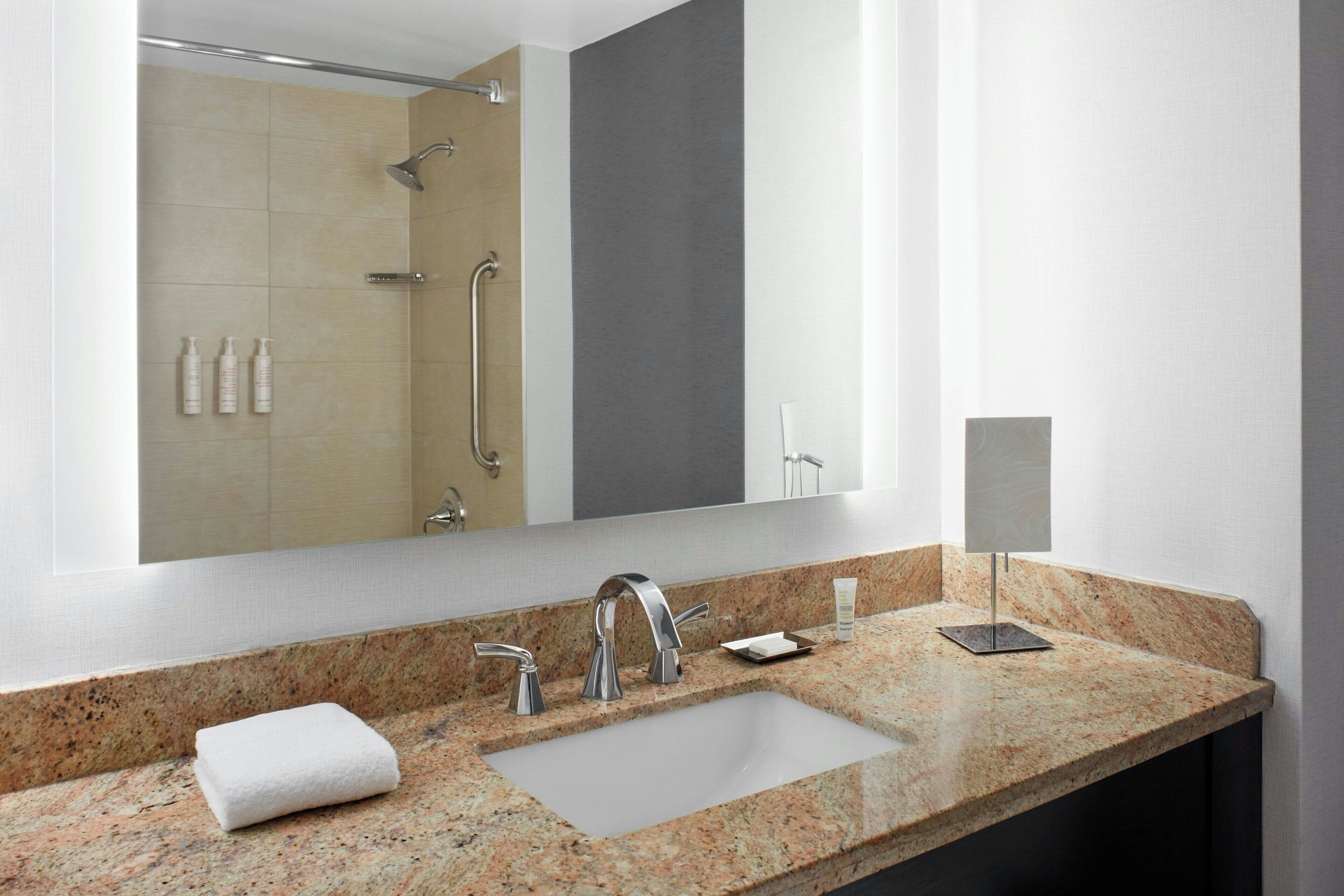 A modern bathroom with a granite countertop, a large mirror, a shower with a handheld showerhead, and a towel on the sink.
