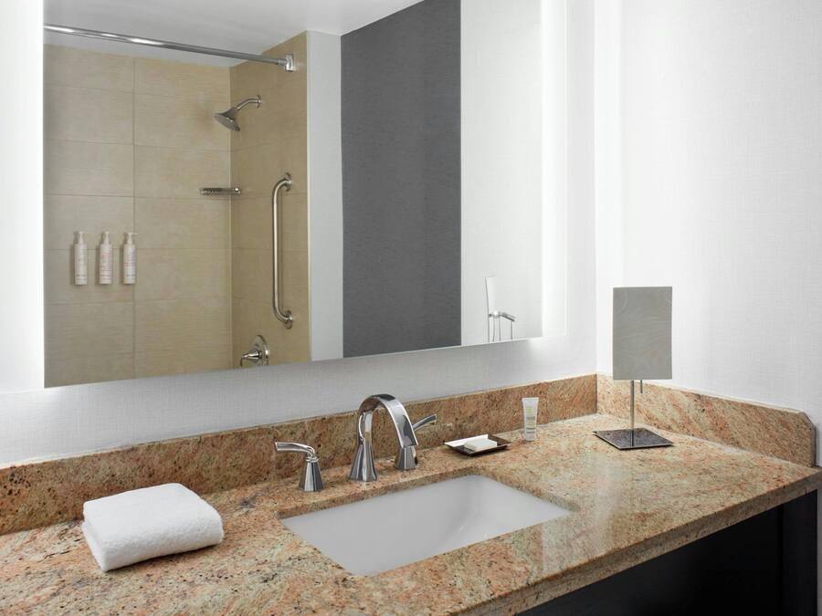 Une salle de bain moderne avec un comptoir en granit, un grand miroir, une douche équipée d’un pommeau de douche à main et une serviette posée sur le lavabo.