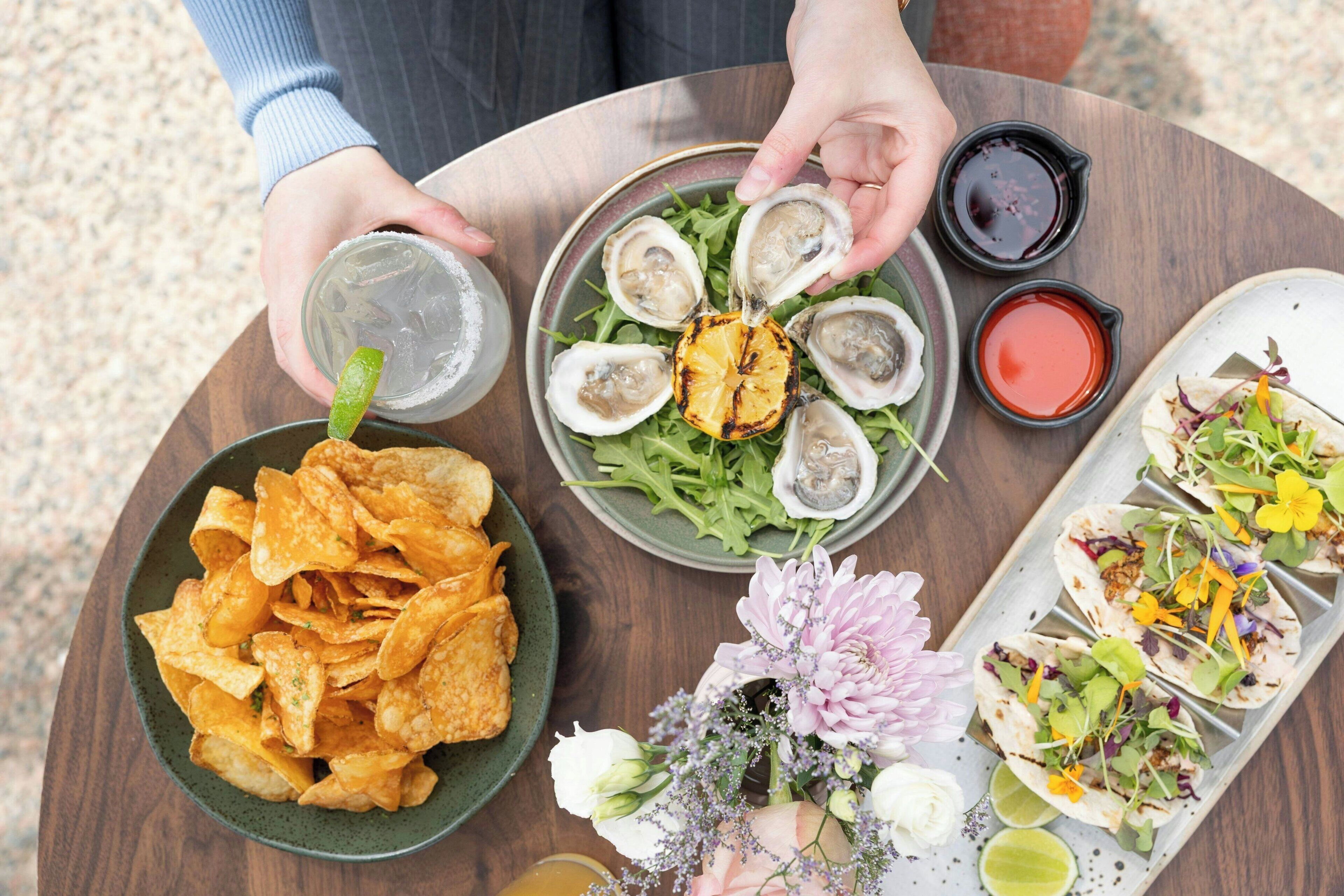 A table with a variety of food items including chips, oysters, and tacos, along with drinks and condiments.