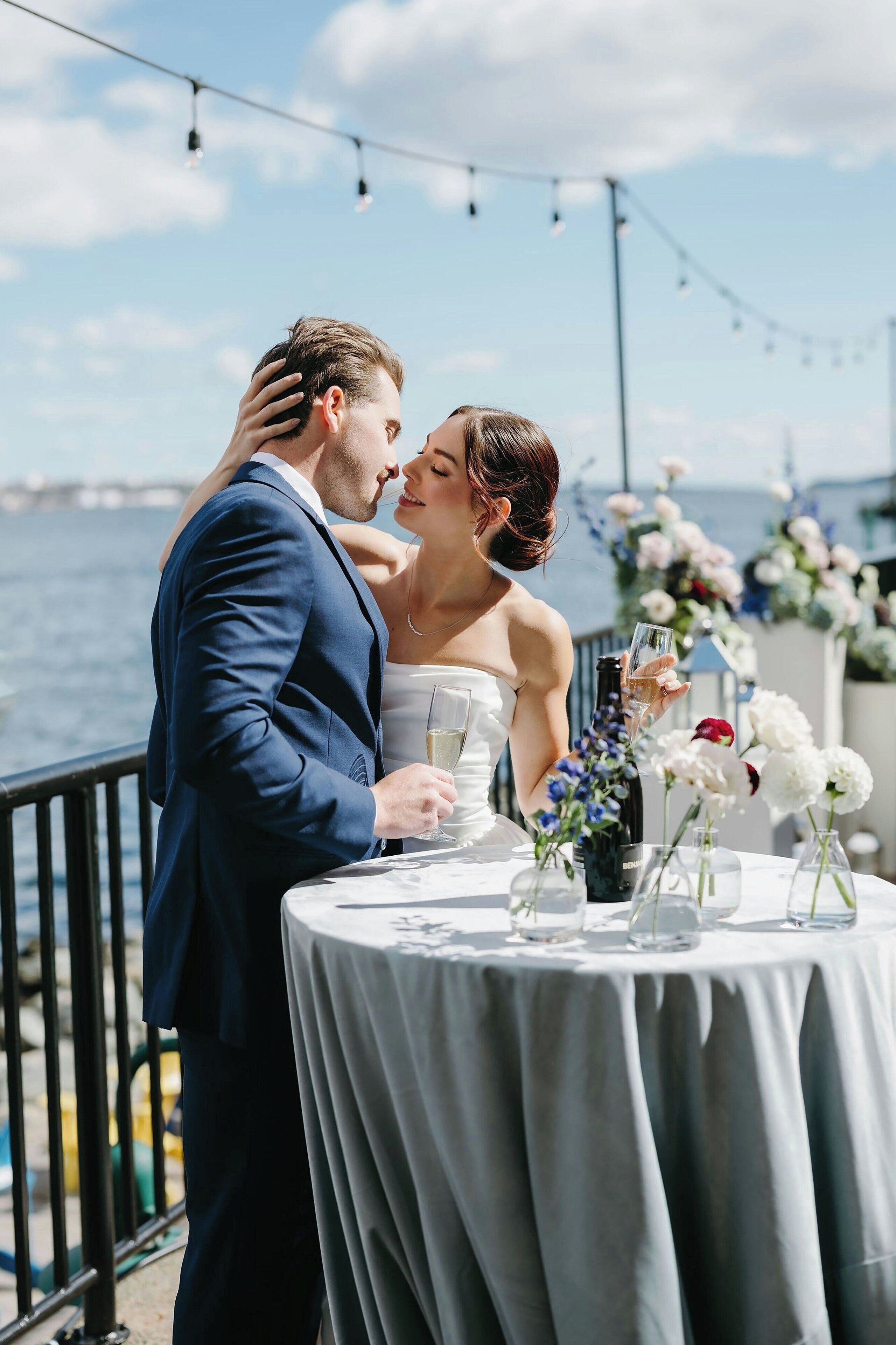 A couple at a table with a view of the sea, a string of lights, and floral decorations.