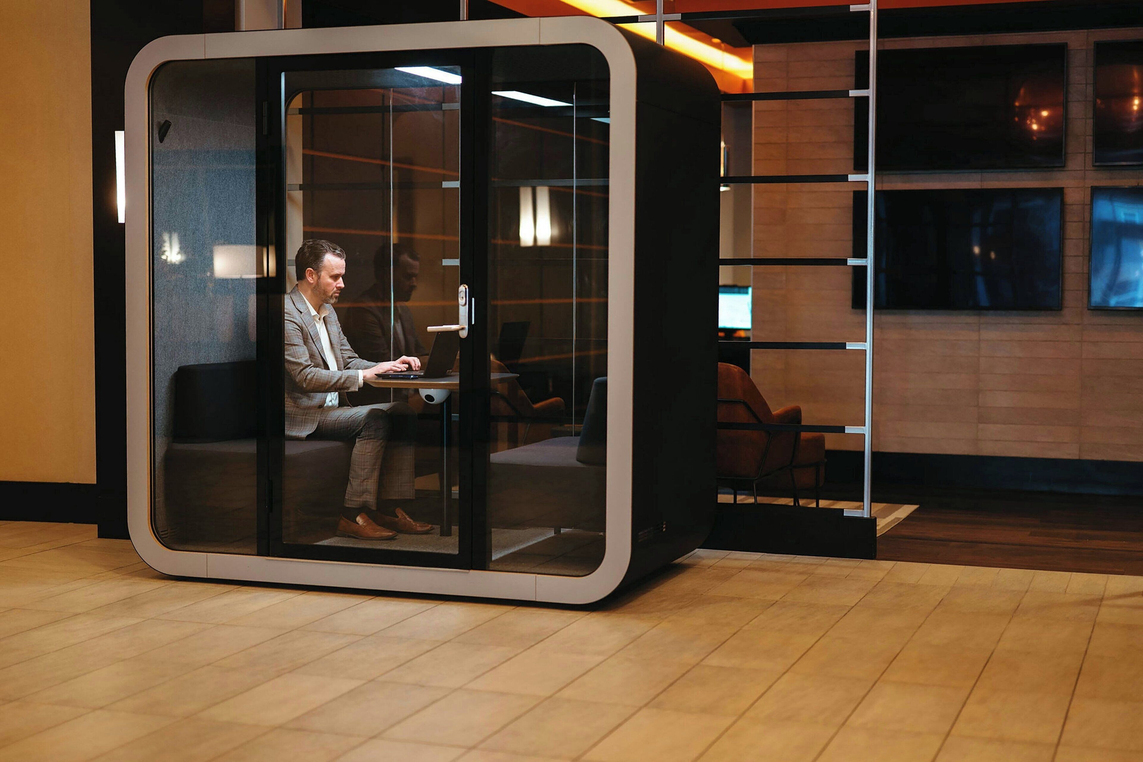 A modern office space with a glass-walled cubicle, a person working at a desk, and a wooden wall with multiple monitors.