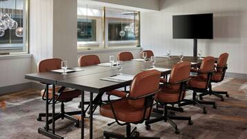 Un salón de conferencias con una larga mesa, ocho sillas de cuero marrón, un televisor de pantalla plana y grandes ventanales con lámparas colgantes decorativas.