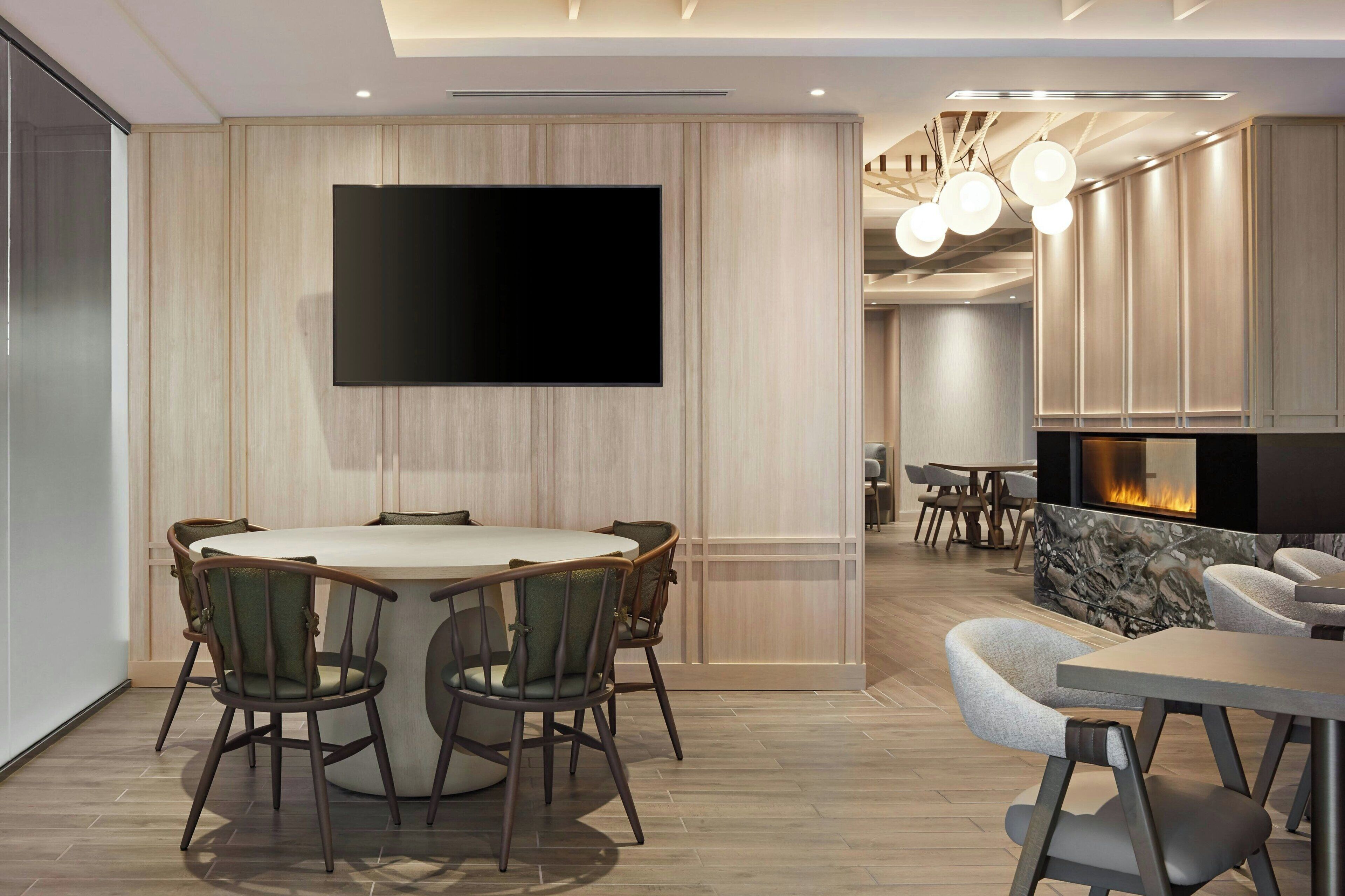 A modern dining area with a round table and chairs, a flat-screen TV mounted on the wall, and a fireplace in the background.